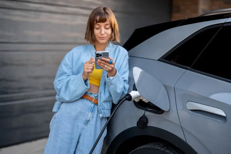 Dekoratives Bild. Eine Frau steht angelehnt ans Auto, hält ein Handy in der Hand. Der Tankdeckel eines Elektroautos ist geöffnet und ist an der Ladesäule angeschlossen. 